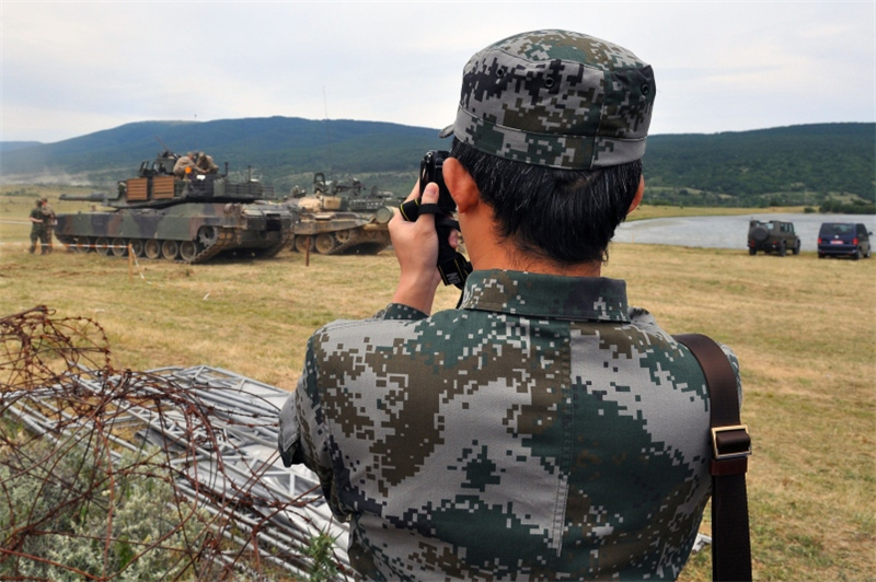 一名来自中国军队的摄影师在现场拍照，其前方是参加此次演习的美军M1A2主战坦克。除了中国外，还有来自波斯尼亚、罗马尼亚和塞尔维亚等国的军事观察员观看了演习。